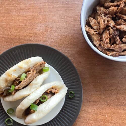 two pulled duck bao on a plate next to a bowl of pulled duck.