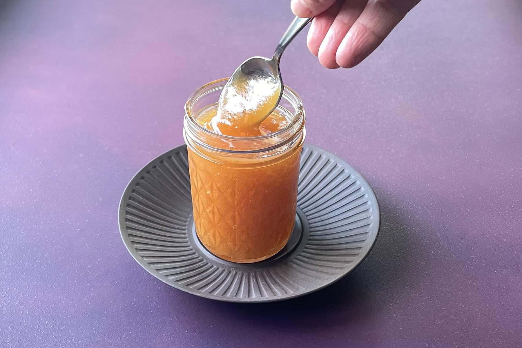 hand dipping spoon into jar of apple curd on a plate.