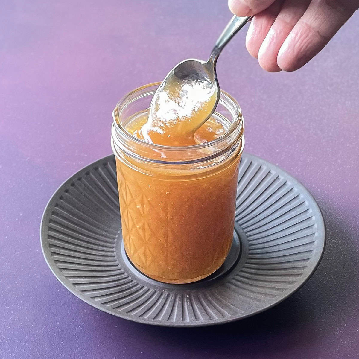 hand dipping spoon into jar of light brown apple curd on a plate.