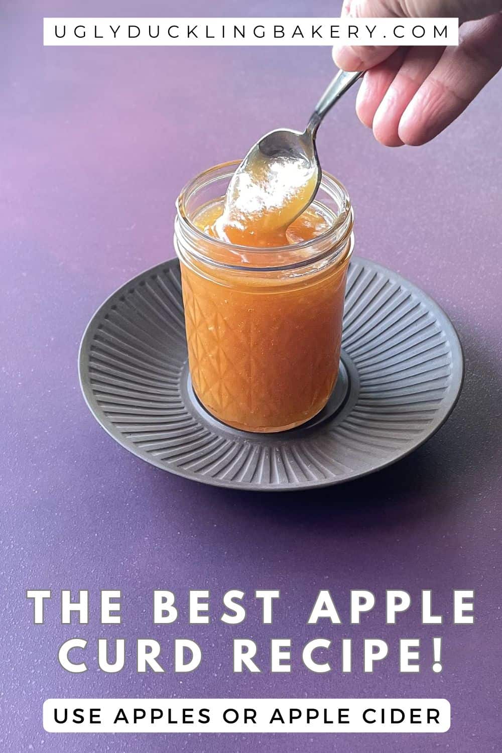 hand dipping a spoon into a glass jar of apple curd. The title says The Best Apple Curd Recipe. Use apples or apple cider. The website ugly duckling bakery dot com is at the top.
