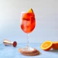wine glass filled with a negroni spritz, ice cubes, and orange slice next to jigger and cut orange.