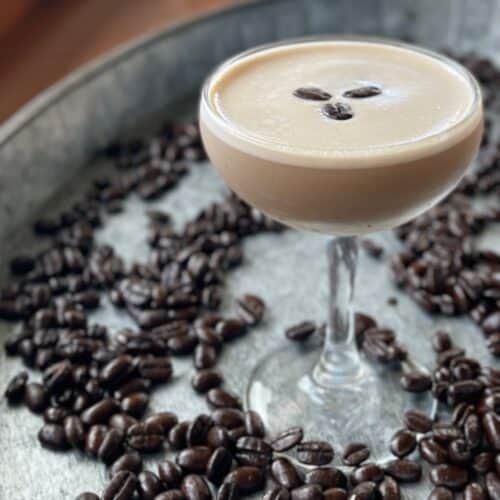 coupe with a light brown cocktail and creamy top layer garnished with three coffee beans on a tray of coffee beans.