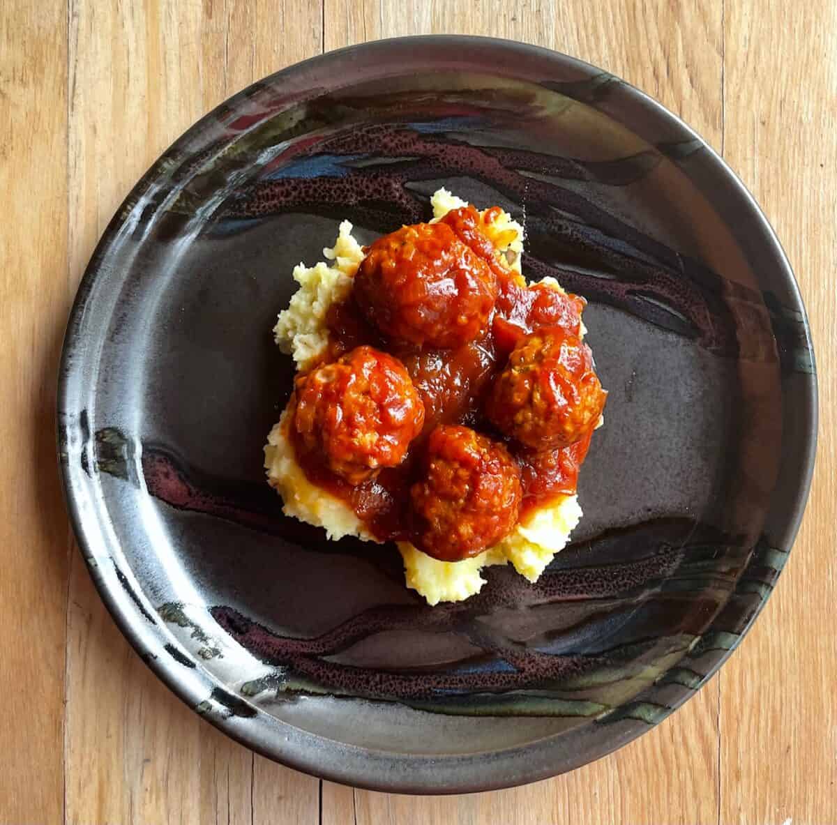 four BBQ meatballs over mashed potatoes on a black plate on a wood table.