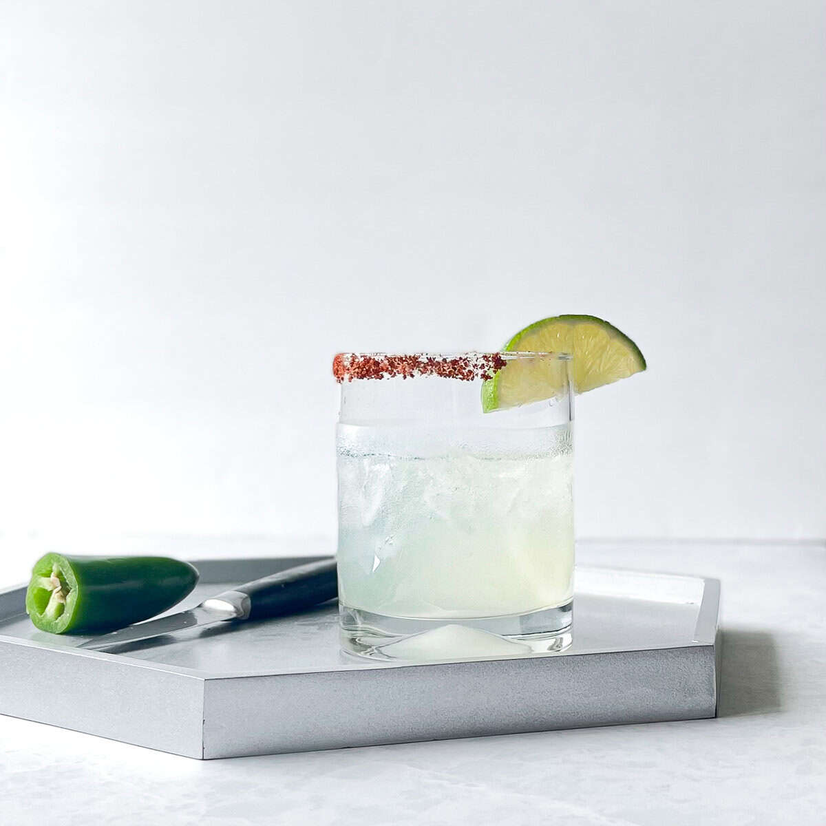 lowball glass filled with crushed ice and pale cocktail. The glass is rimmed with a reddish salt and there is a lime wedge garnish. The cocktail sits on a silver tray with a cut jalapeno and paring knife.