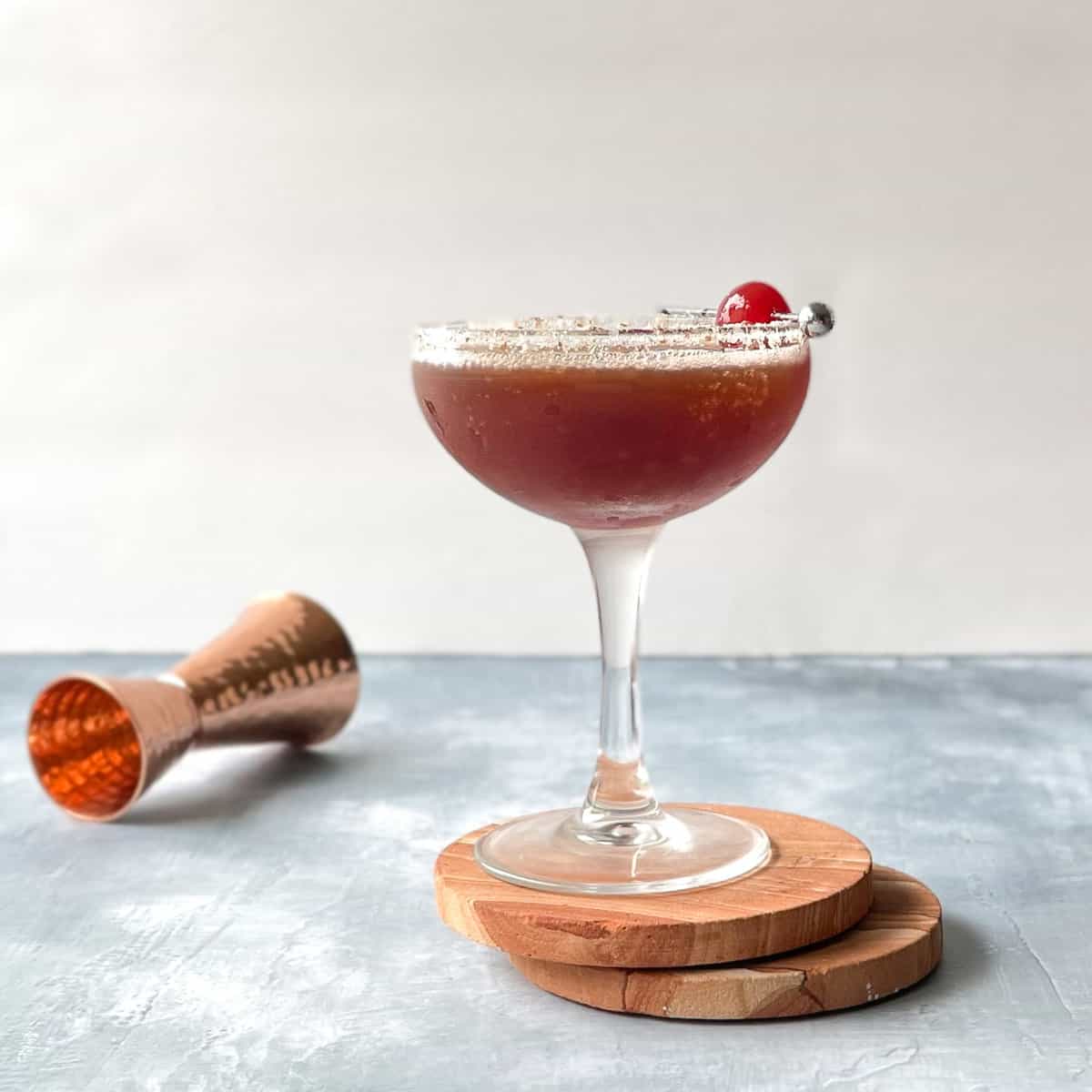 red brown cocktail in an almond-sugar rimmed coupe with a maraschino cherry garnish.