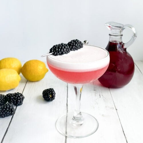 pink cocktail with egg white foam and blackberry garnish with small carafe of purplish red syrup, lemons, and blackberries in background.