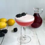 a pink cocktail with foam and blackberry garnish with small carafe of purplish red syrup, lemons, and blackberries in background.