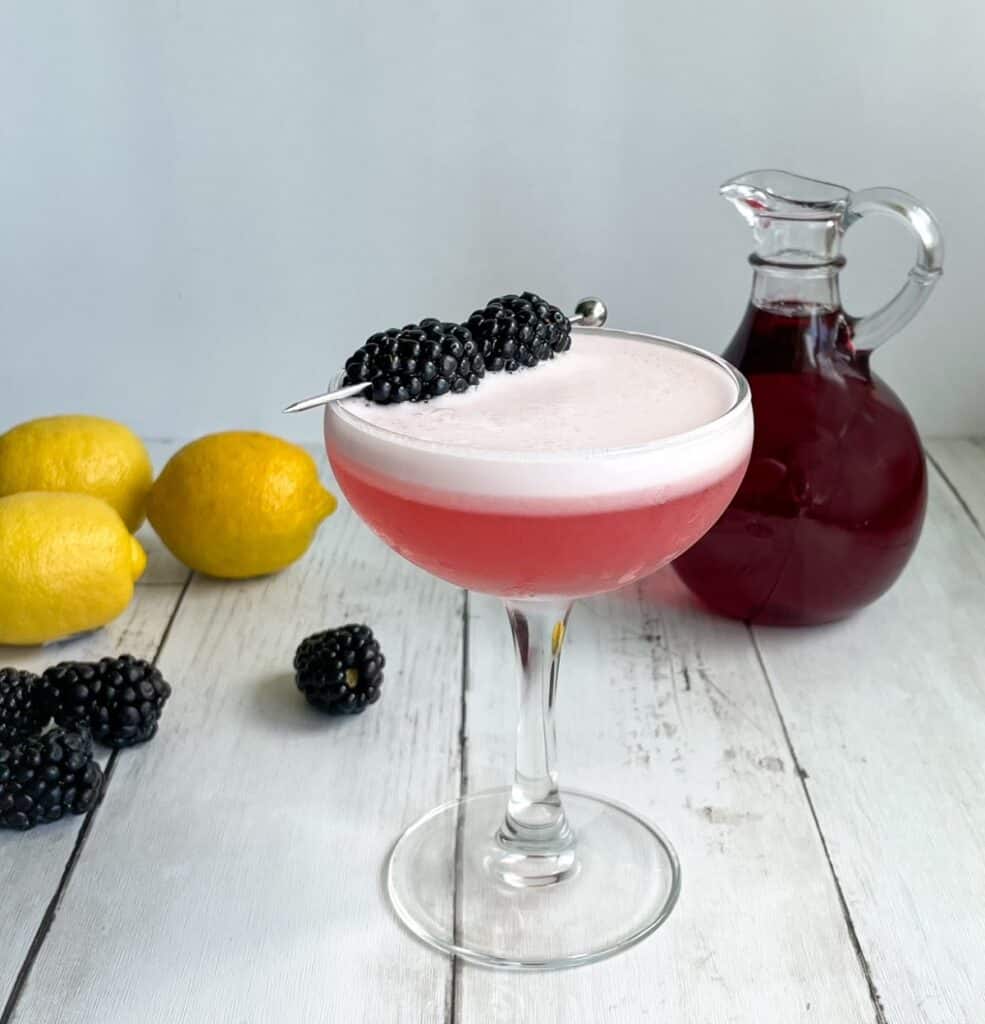 a pink vodka sour cocktail with foam and blackberry garnish with blackberry simple syrup, lemons, and blackberries.