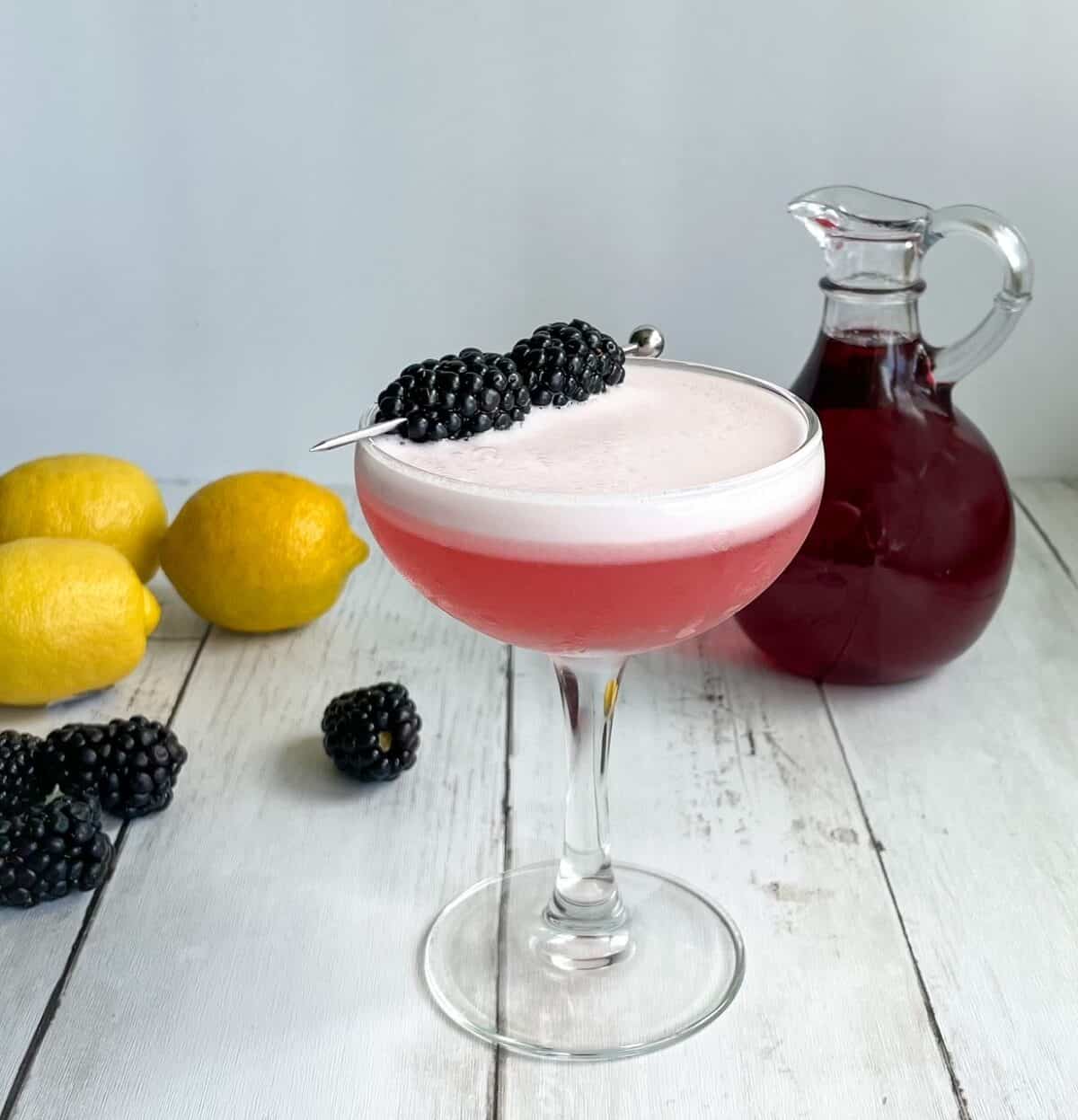 a pink cocktail with foam and blackberry garnish with small carafe of purplish red syrup, lemons, and blackberries in background.