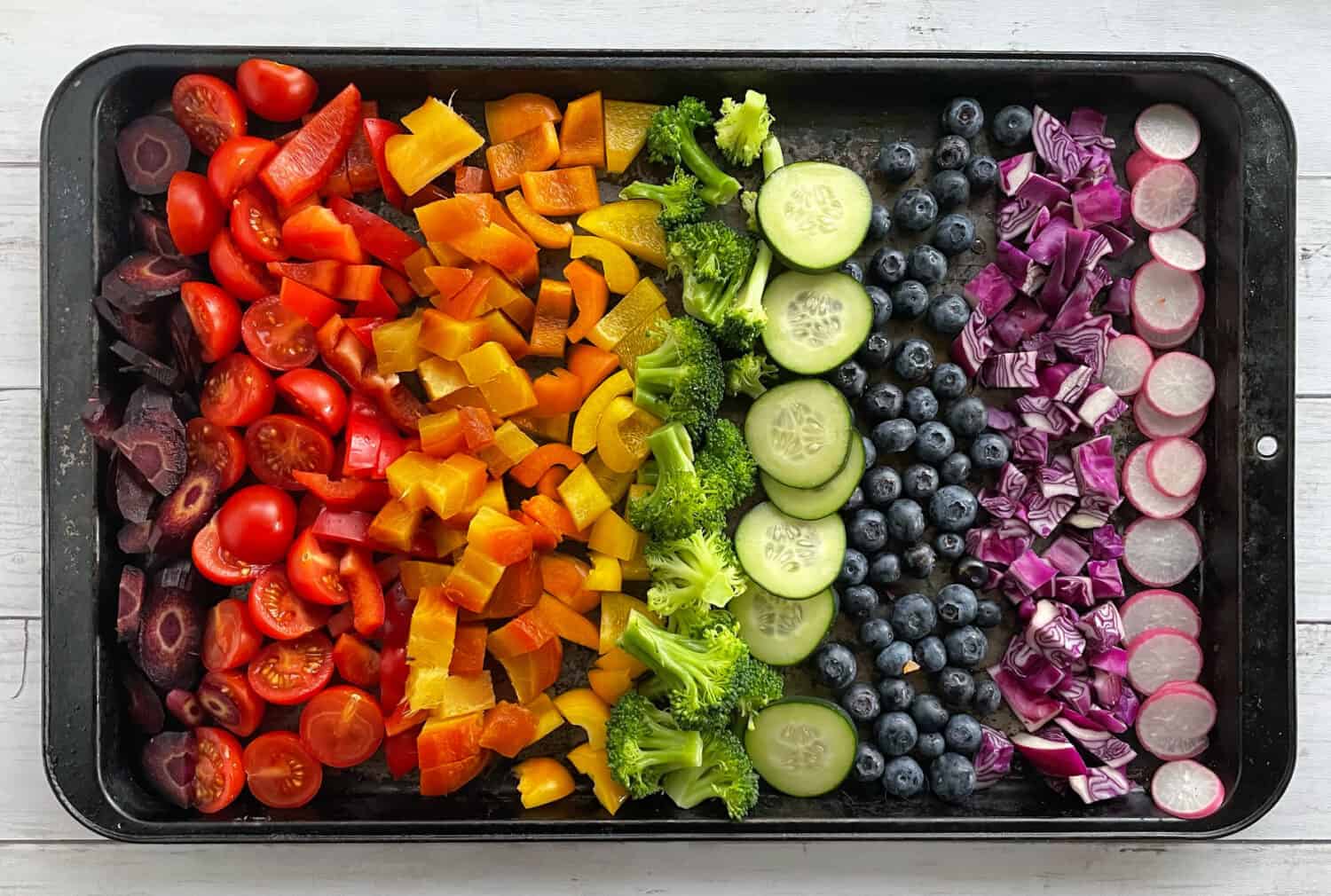 cut vegetables on a baking sheet in rainbow order including purple carrots, tomatoes, red pepper, chickpeas, orange and yellow pepper, broccoli, blueberries, red onion, and radishes.