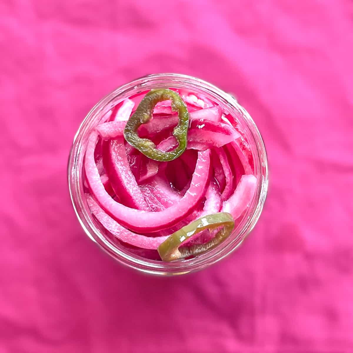 a glass jar of pink pickled onions with sliced jalapeno peppers.