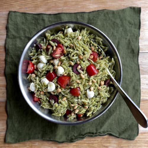 a bowl of orzo pesto salad with cherry tomatoes, mozzarella, olives, and toasted pine nuts.