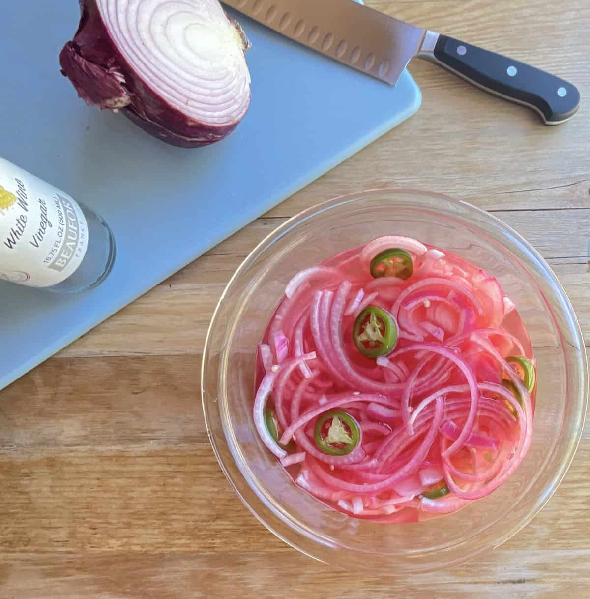 a bowl of pink pickled onions with jalapeno slices next to a cutting board with onion half, knife, and vinegar bottle.