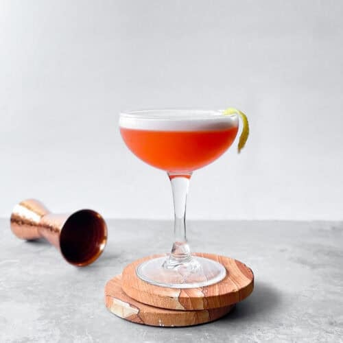 coupe with a bright orange cocktail with foam and lemon twist garnish on two coasters and next to a brass jigger.