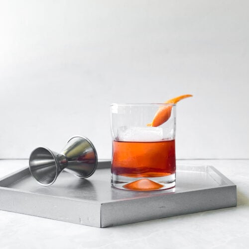 orange cocktail in a low ball glass with large ice cube and orange twist. It is on a grey tray with a silver jigger.
