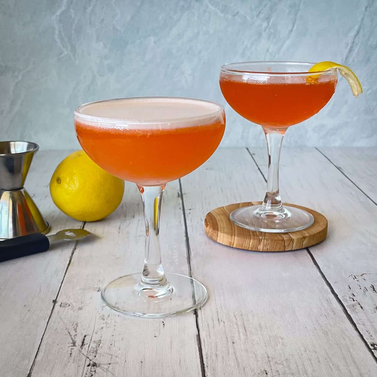 two bright orange cocktails. The one in front has an egg white foam. The one in back is on a coaster and has a lemon twist. There is also a lemon, zester, and jigger.