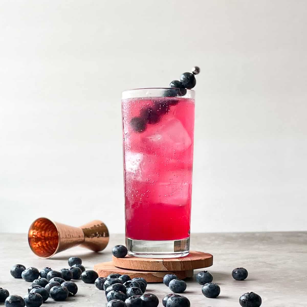 deep pink cocktail in highball glass with ice surrounded by fresh blueberries.