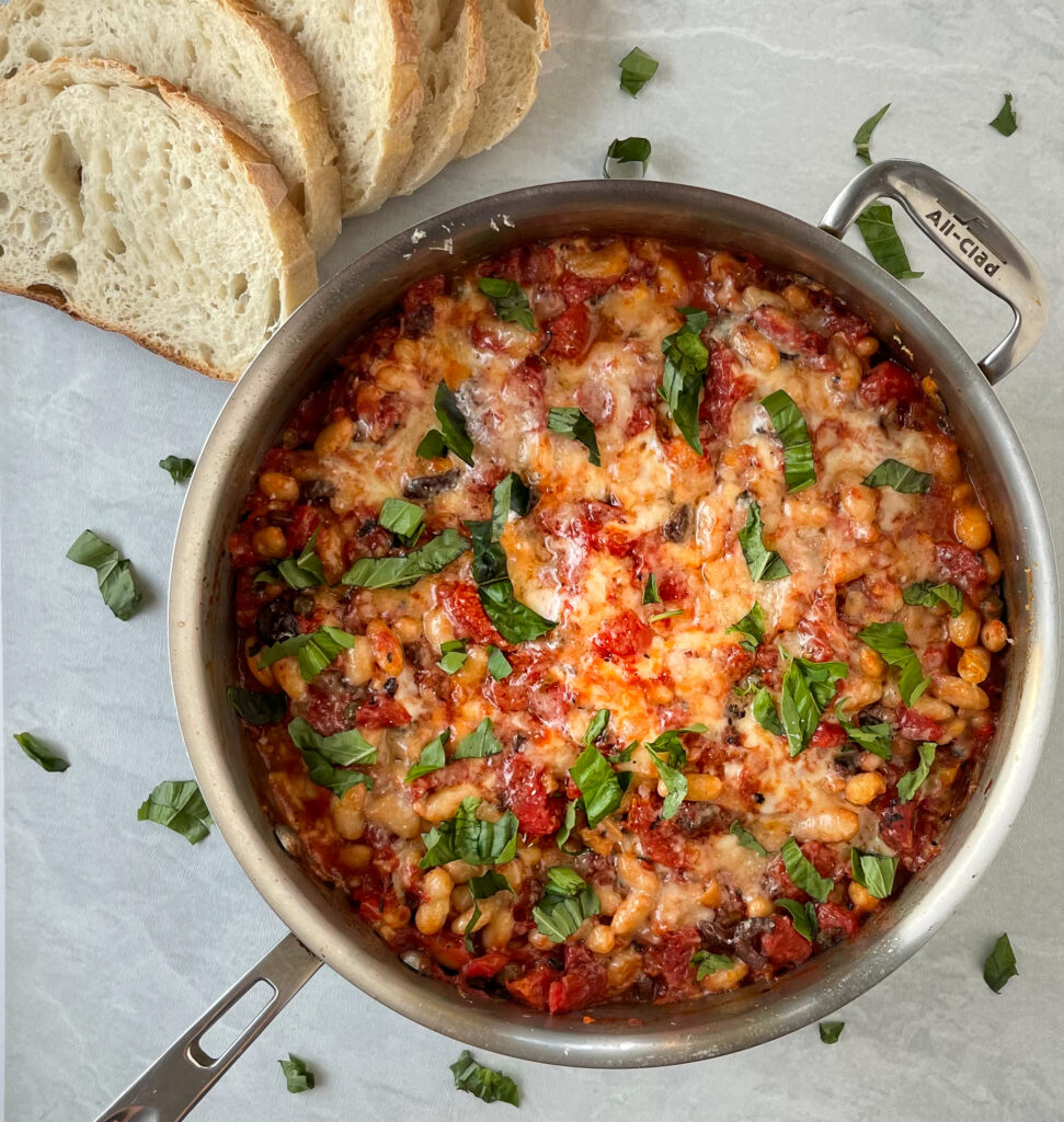 a large saute pan filled with tomatoes, white beans, cheese, and sprinkled with basil next to a few slices of crusty white bread.