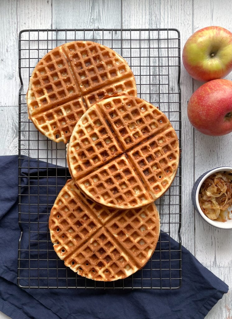 three waffles on a cooling rack next to two apples and a small bowl of caramelized onions.