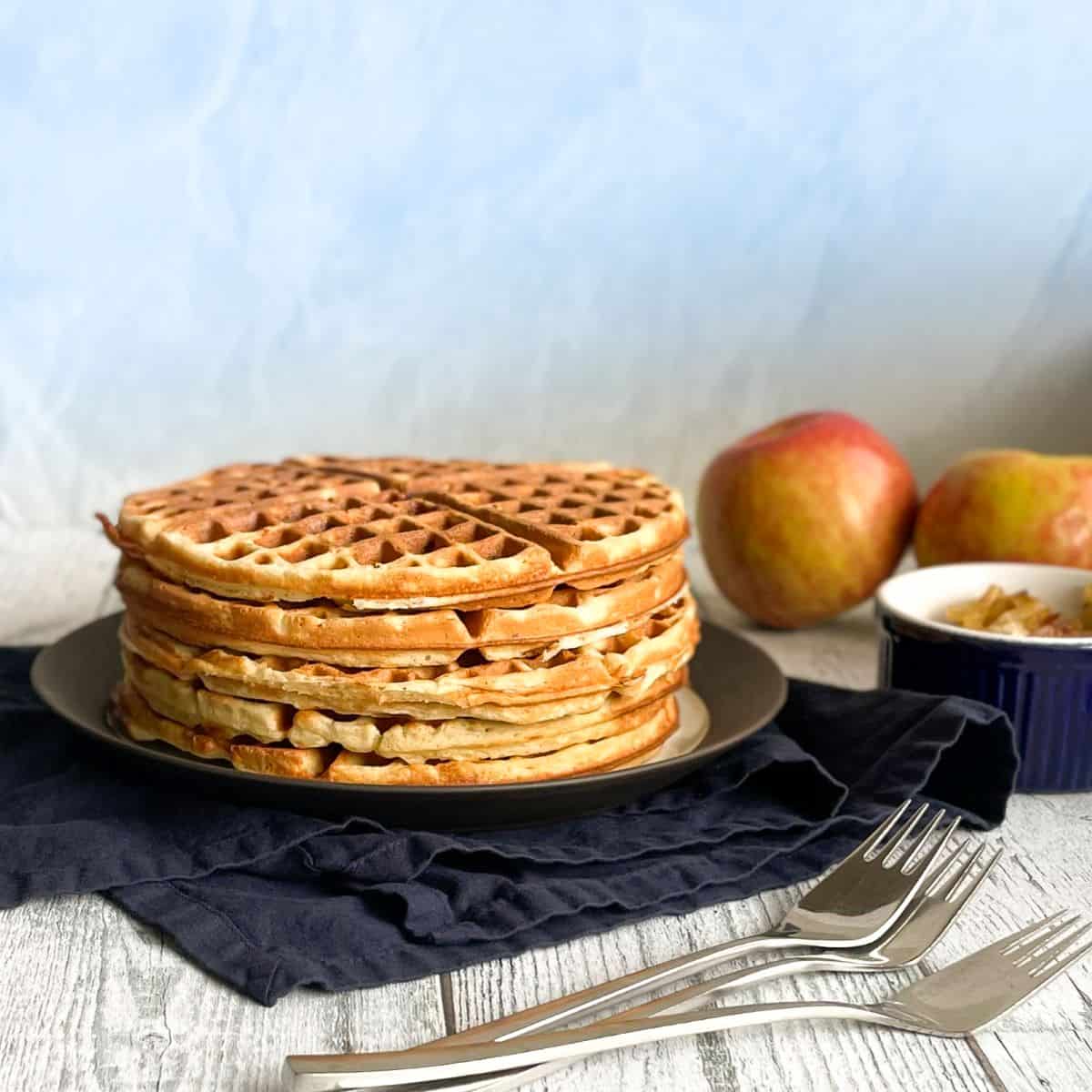 a stack of apple waffles on a plate with three forks, two apples, and caramelized onion bowl.