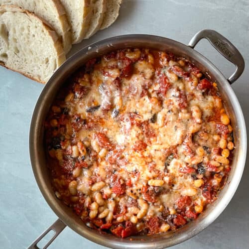 a pan full of cheesy beans next to slices of crusty bread.