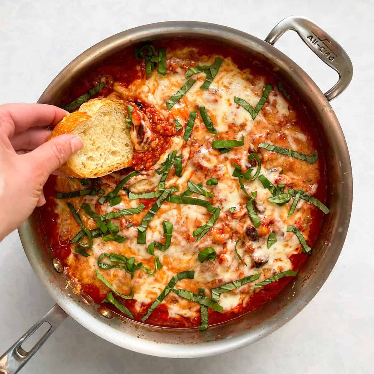 frying pan with a hand dipping a piece of crusty toast into frying pan full of beans, tomato sauce, and melted cheese topped with basil slivers.