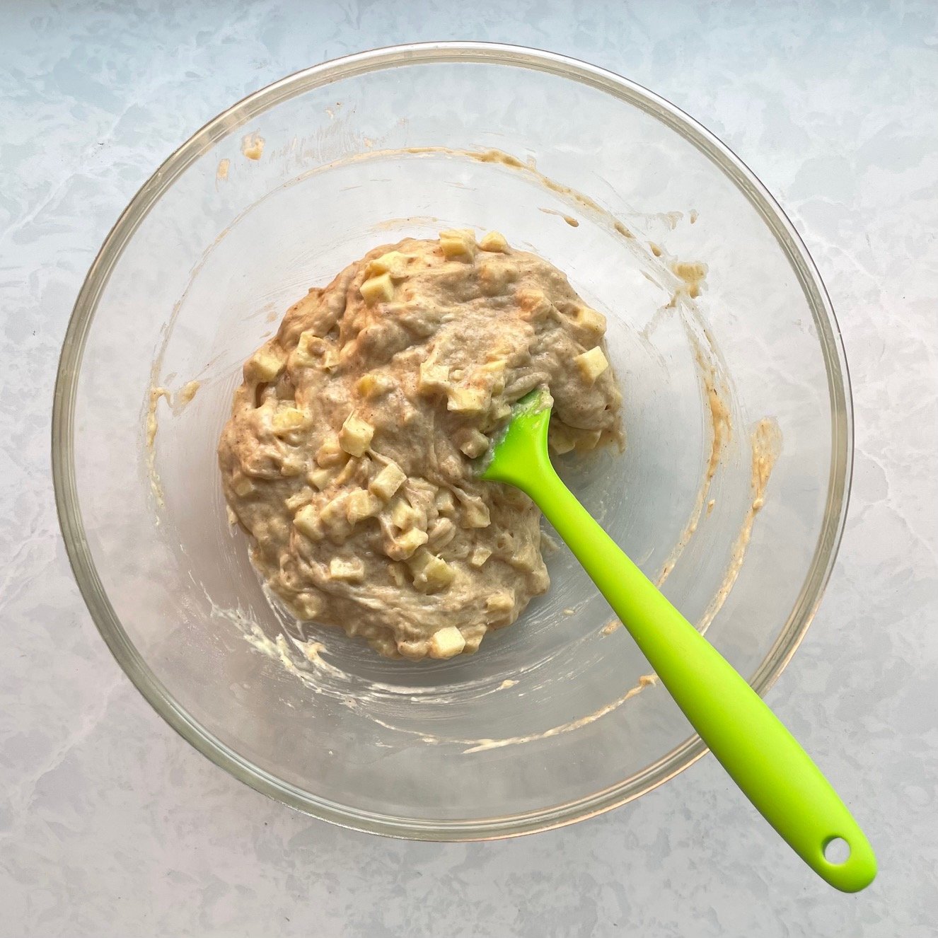 light brown batter with apple chunks in a large glass bowl with green silicone spatula.