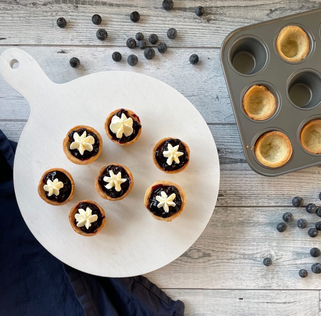seven miniature blueberry pies topped with whipped cream surrounded by more mini pie shells and fresh blueberries.