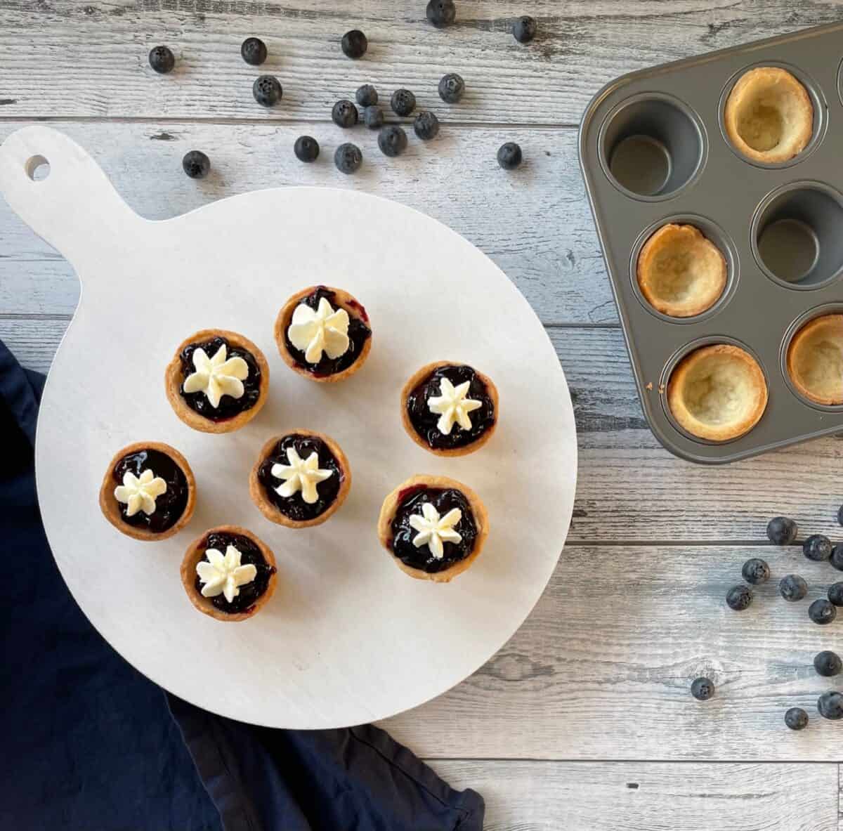 seven miniature blueberry pies topped with whipped cream surrounded by more mini pie shells in a mini muffin tin and fresh blueberries scattered on a countertop.