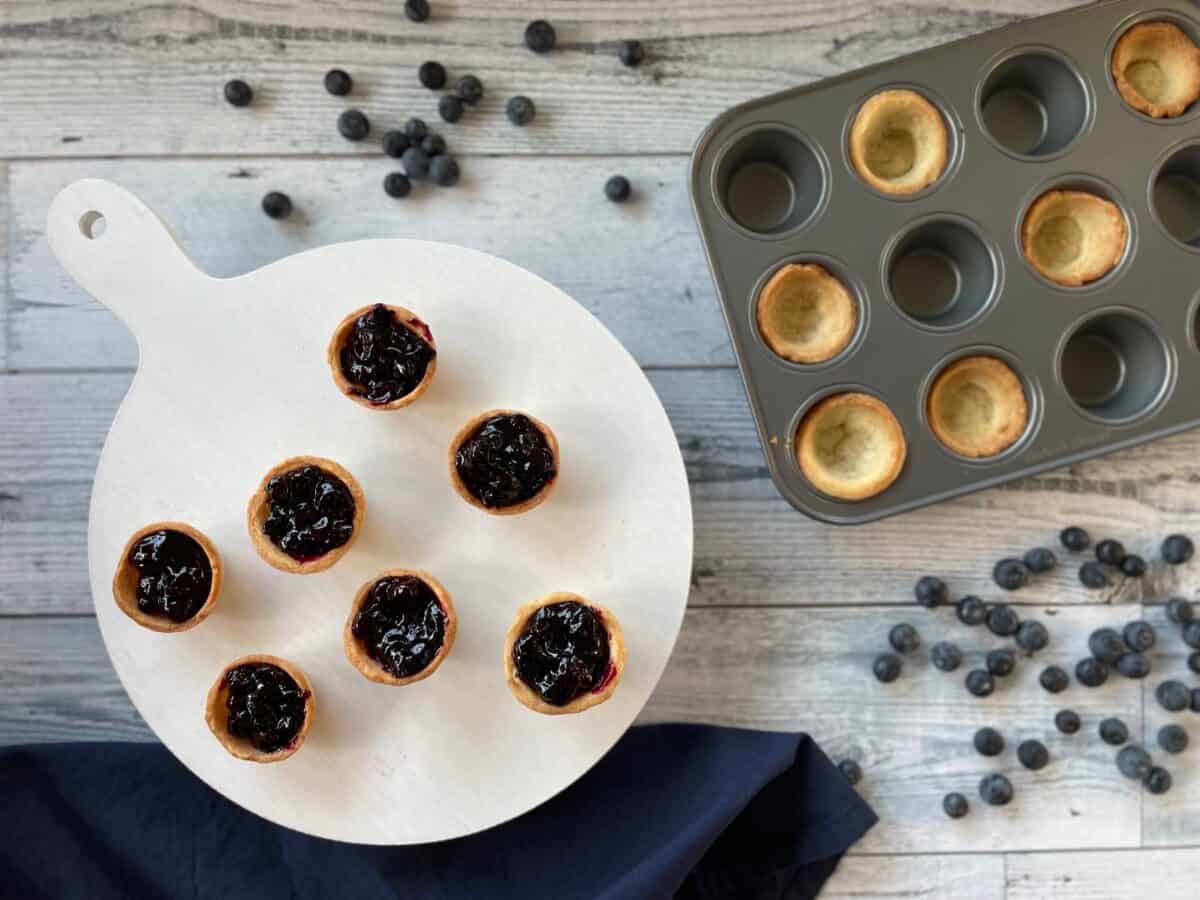 mini blueberry pies on a white serving tray with mini pie shells in a mini muffin tin and fresh blueberries scattered about.