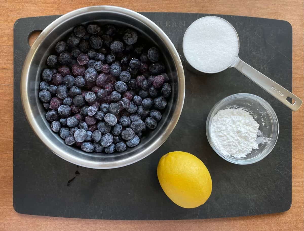 blueberries, sugar, cornstarch, and a lemon on a black cutting board.