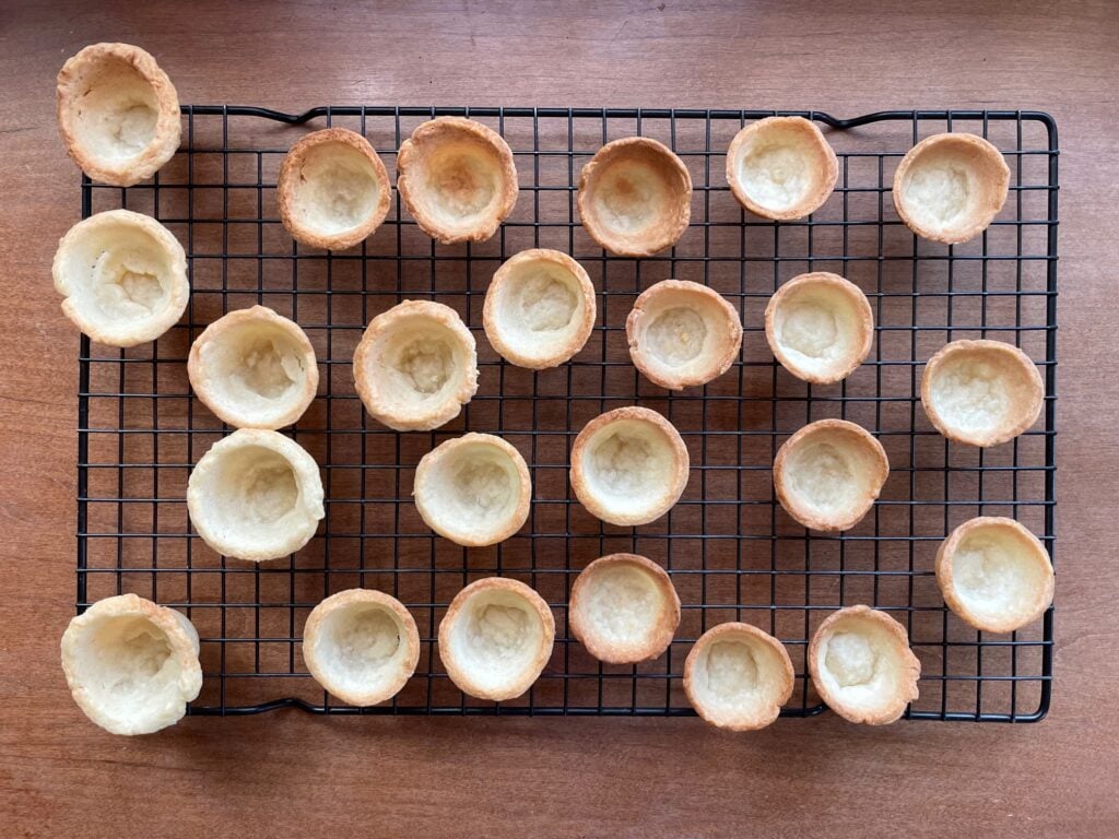 Twenty four baked and empty mini pie crust shells on a cooling rack.