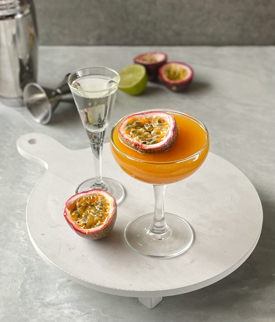 a passion fruit martini on a tray with shot of sparkling wine, passion fruit half, with a cocktail shaker and fruit in background.