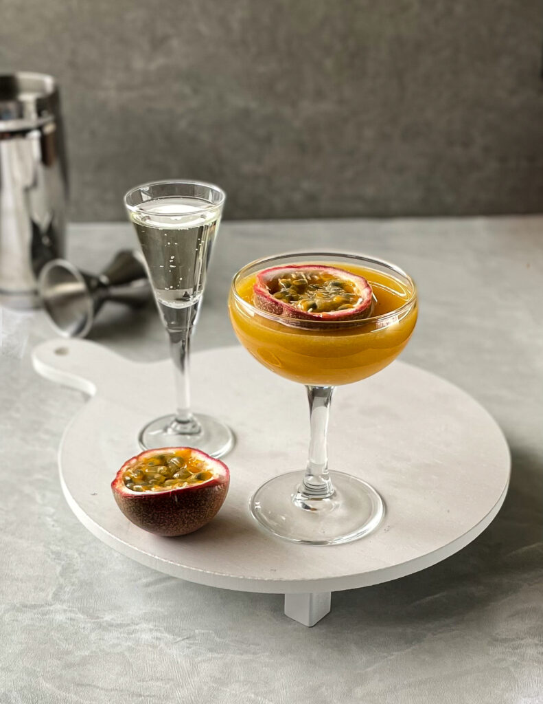 a porn star martini mocktail with a passion fruit garnish on a white tray holding another passion fruit half and glass of bubbly.