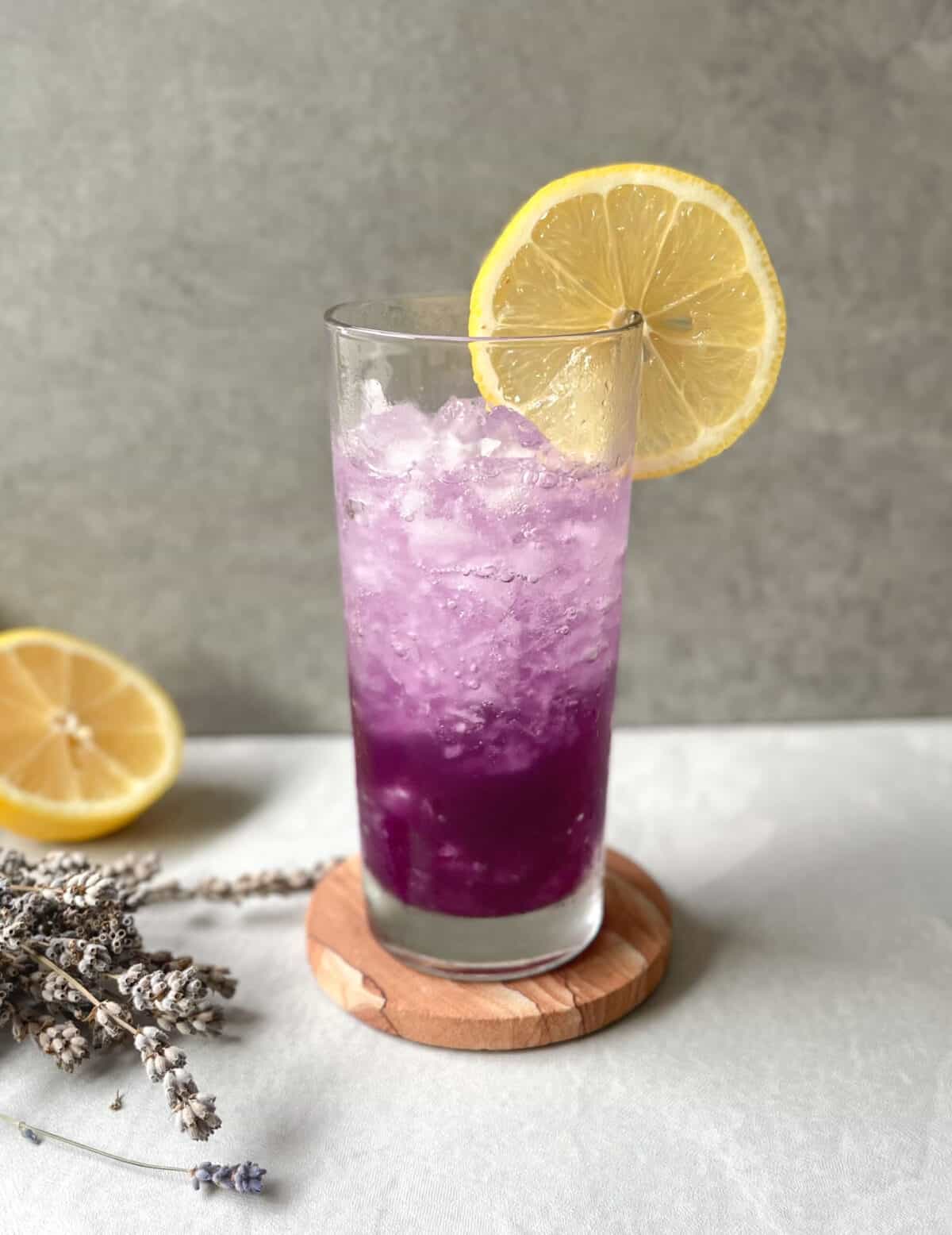 purple cocktail in highball glass garnished with lemon, surrounded by lavender.