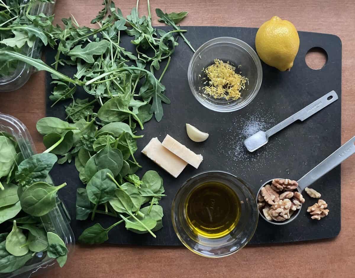 arugula, spinach, garlic, lemon, walnuts, olive oil, and salt on black cutting board.