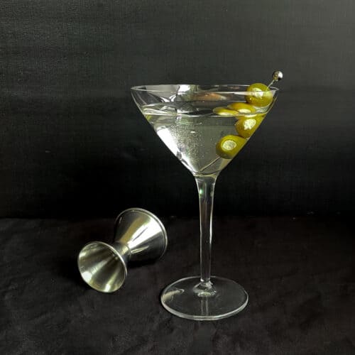 a martini with three blue cheese olives and jigger on a black background.