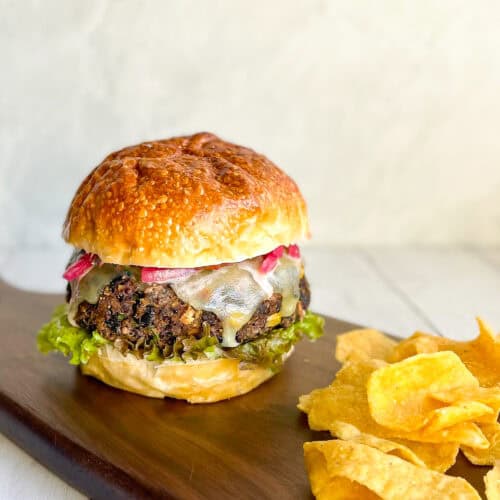 black bean burger on a bun with melting cheese, lettuce, and pickled onions on a wood serving tray with tortilla chips.