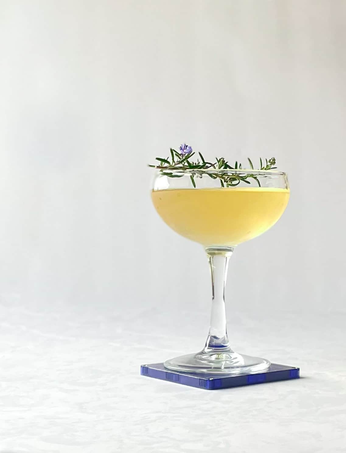 yellow liquid in a coupe garnished with rosemary and on a blue square coaster.