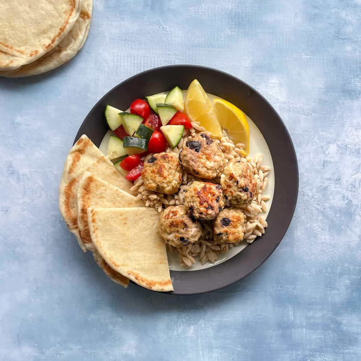 ground chicken meatballs with olives on a plate with pita, orzo, and a tomato and cucumber salad.