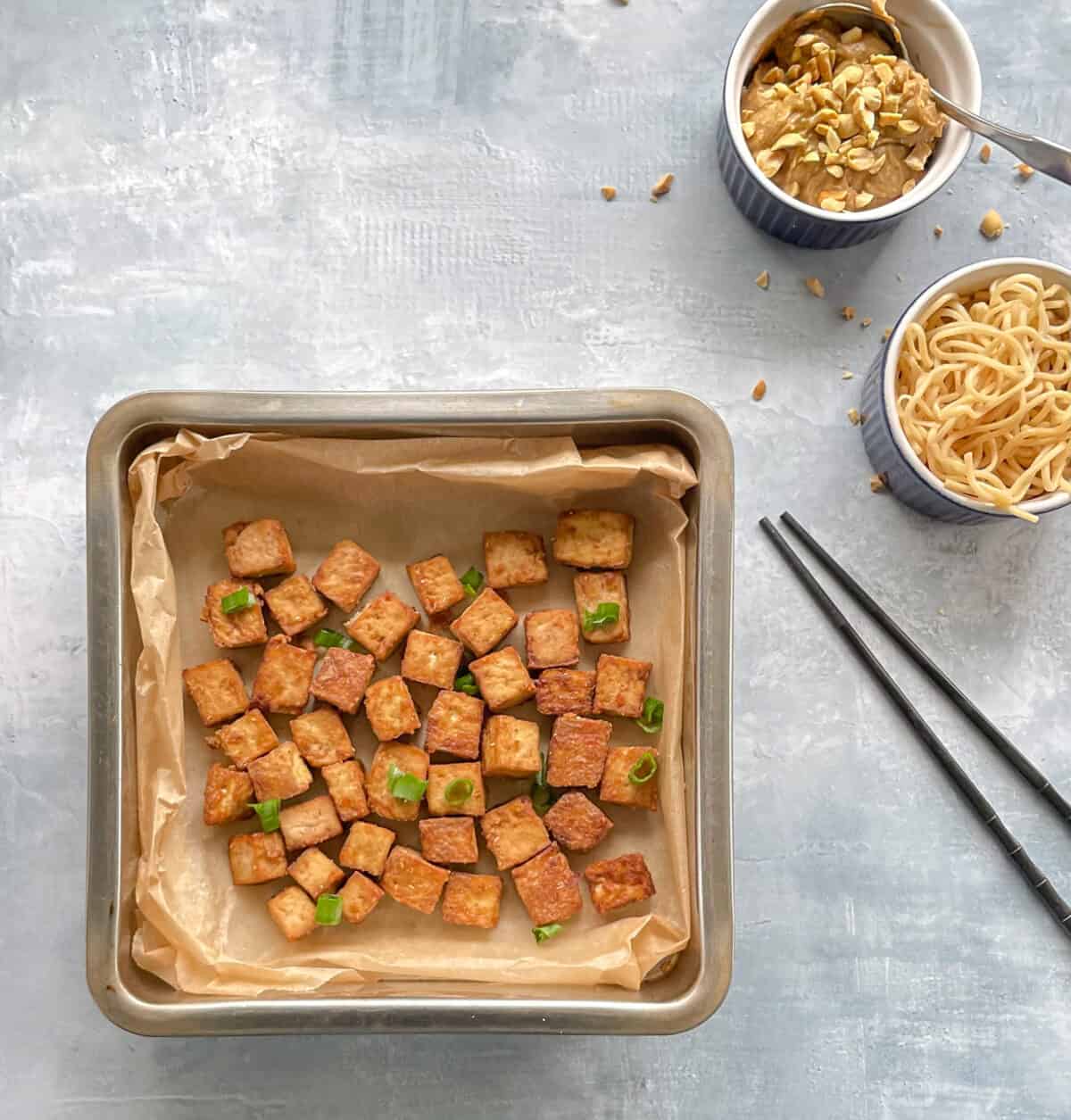parchment-lined pan of crispy baked tofu with noodle bowl, peanut sauce, and chopsticks.