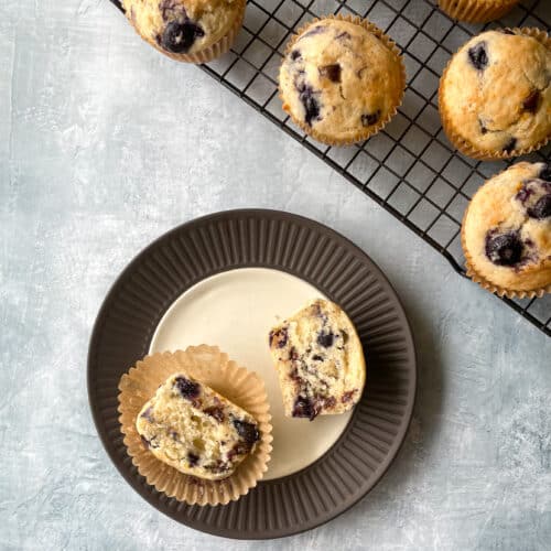 blueberry chocolate chip muffin split open on a plate next to rack of muffins.