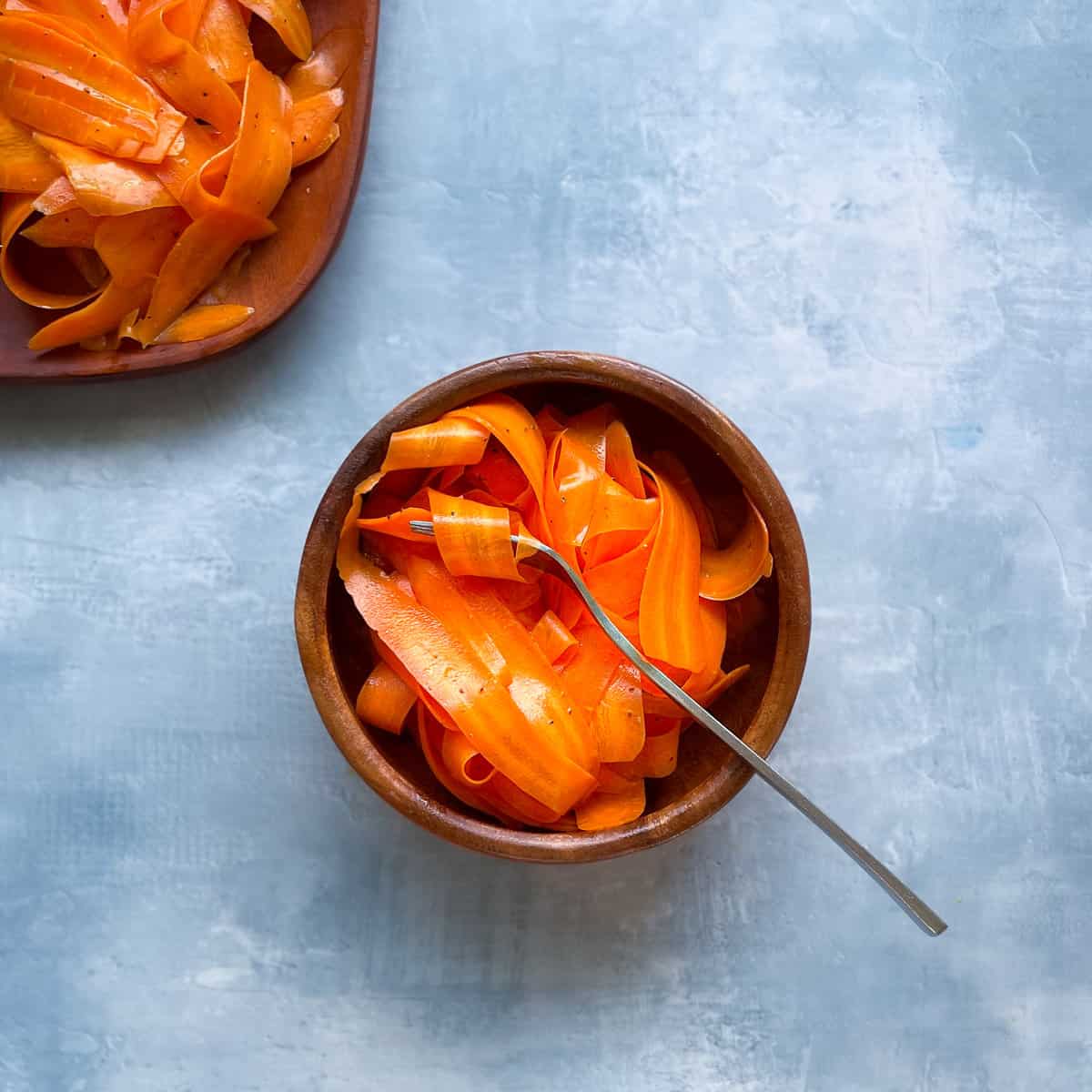 fork and small bowl of strips of raw carrot salad with a larger bowl of carrots in the corner.