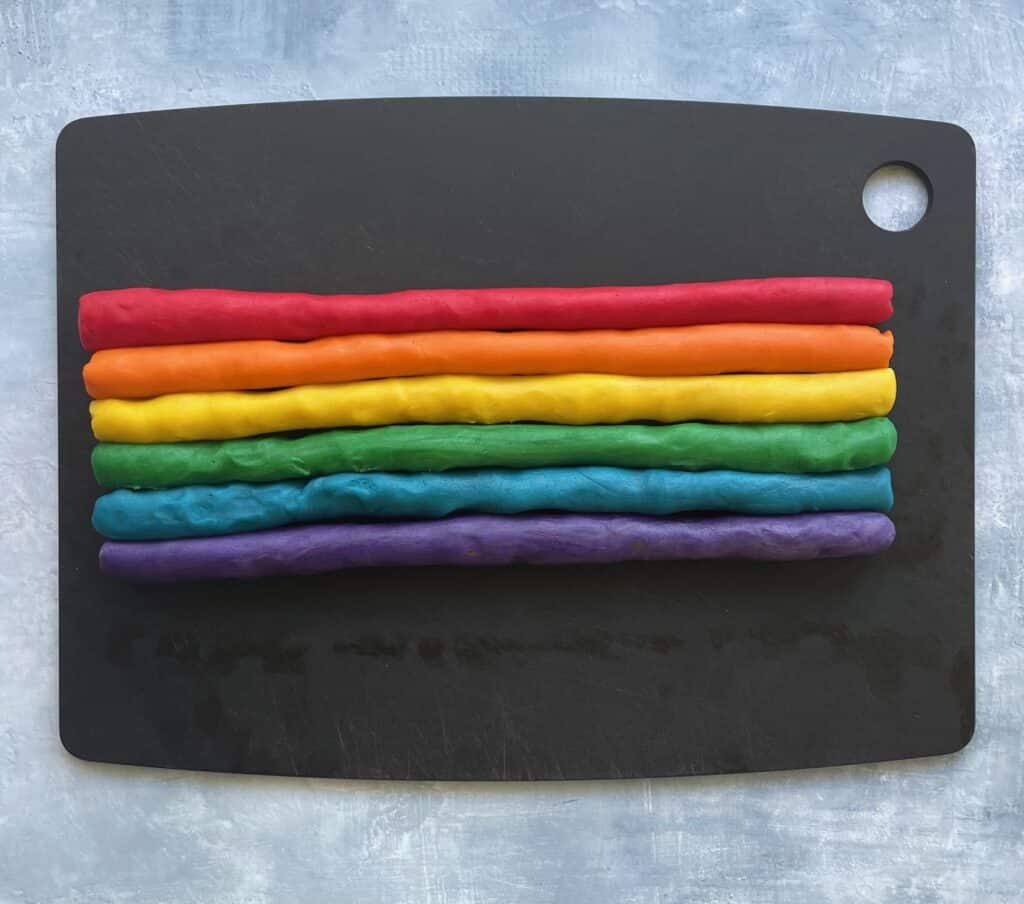 six rolled logs of rainbow colored cookie dough on a cutting board.