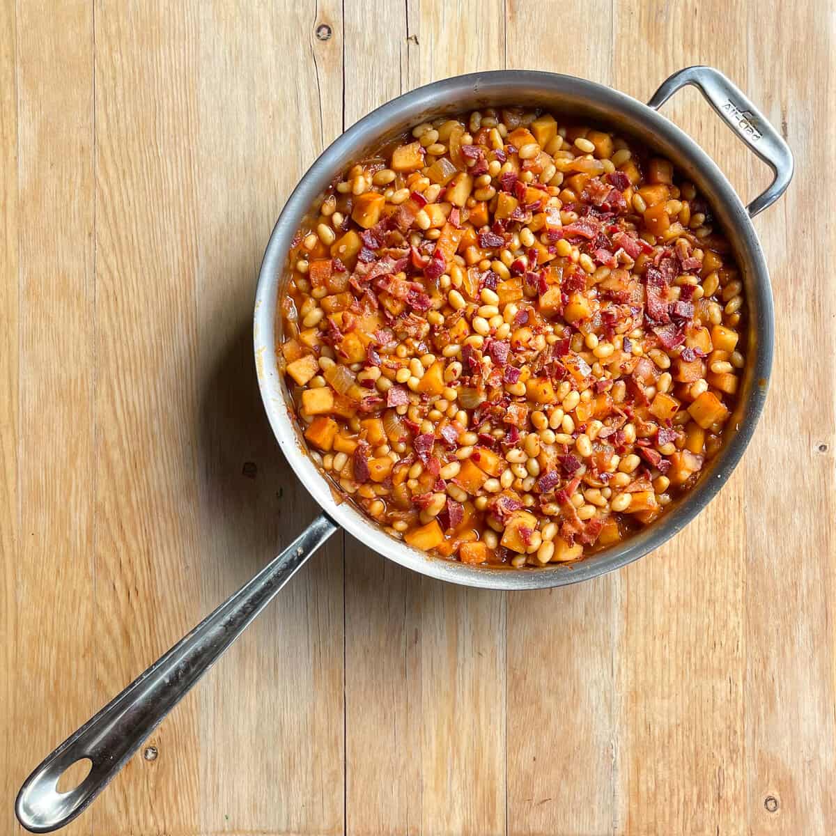 pan of apple pie baked beans topped with bacon on a wood background.