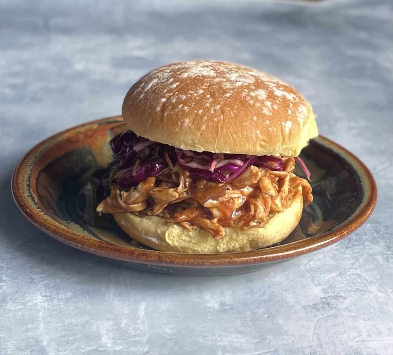 BBQ pulled chicken sandwich with shredded chicken and coleslaw on a bun on a plate.