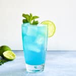 a bright blue mojito cocktail in a highball glass garnished with mint and lime.
