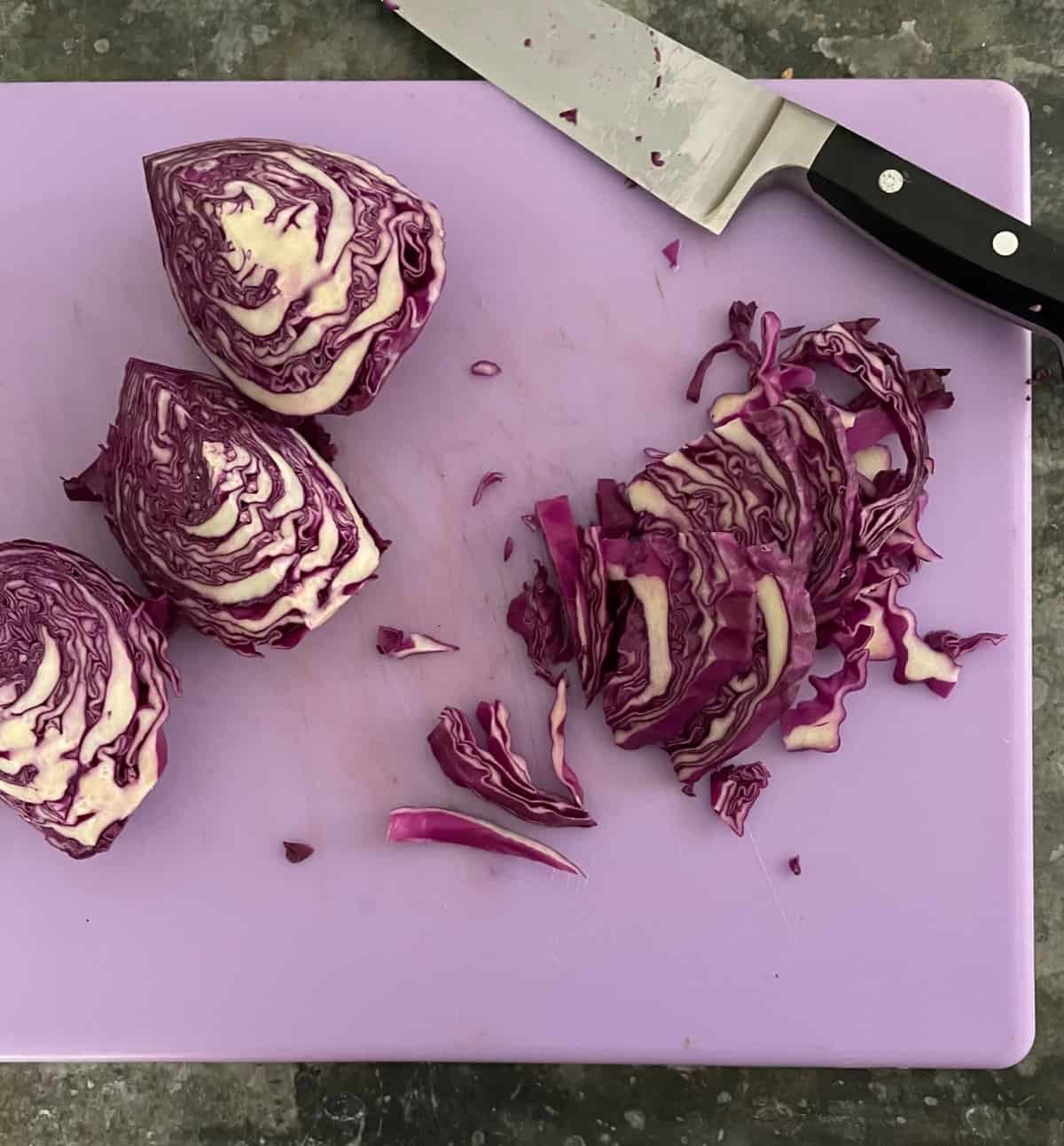 three whole quarters of a purple cabbage, shredded cabbage, and a chef's knife on a purple cutting board.