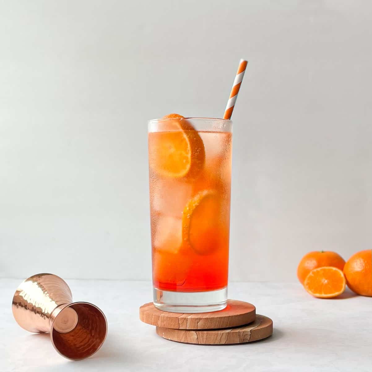 orange highball cocktail with orange slices, ice cubes, and orange striped straw.