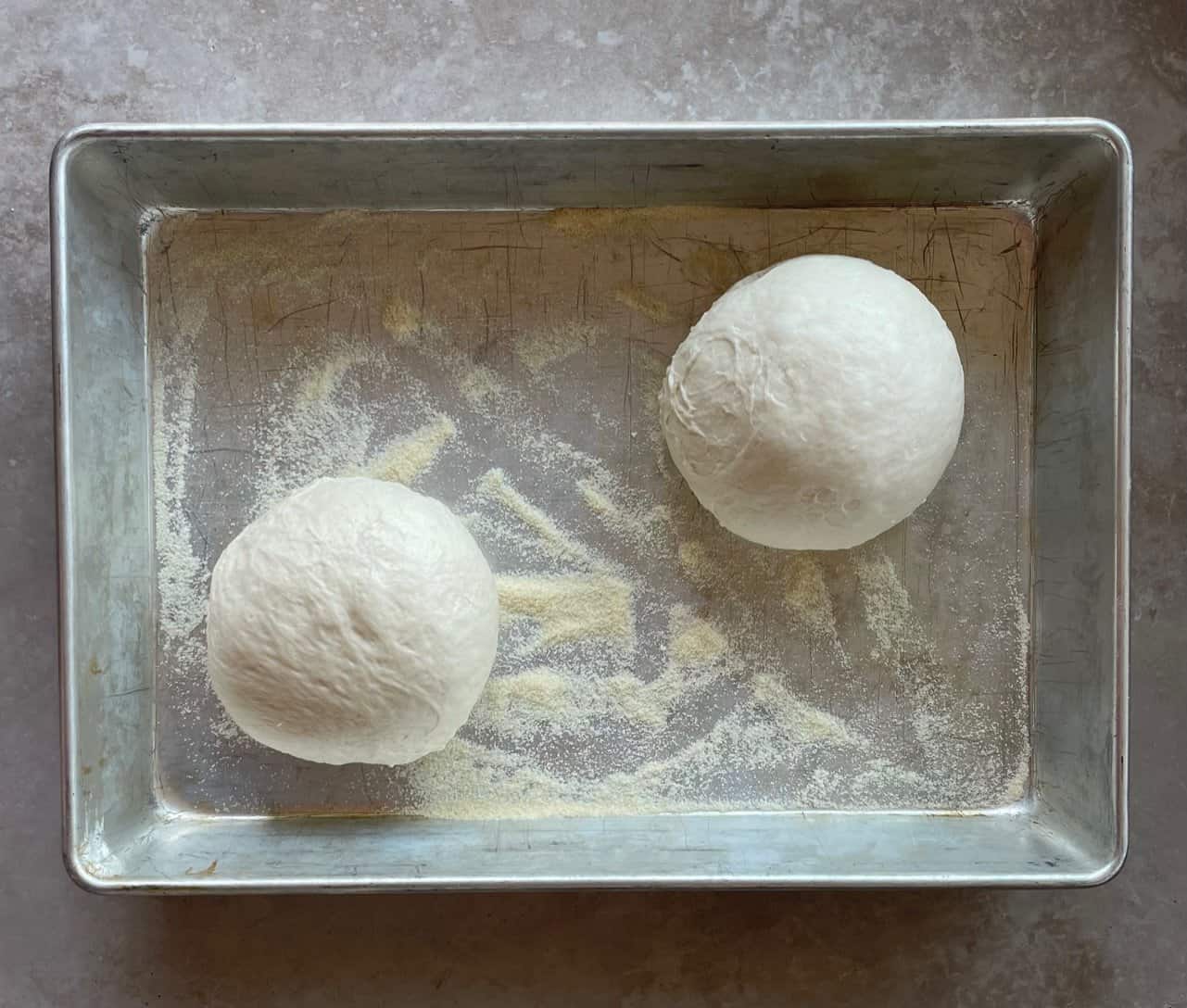 two balls of pizza dough rising in a floured baking pan.